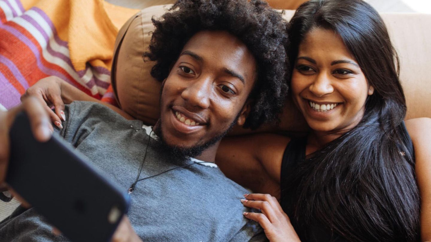 A happy couple lounging together, smiling while watching something on a smartphone