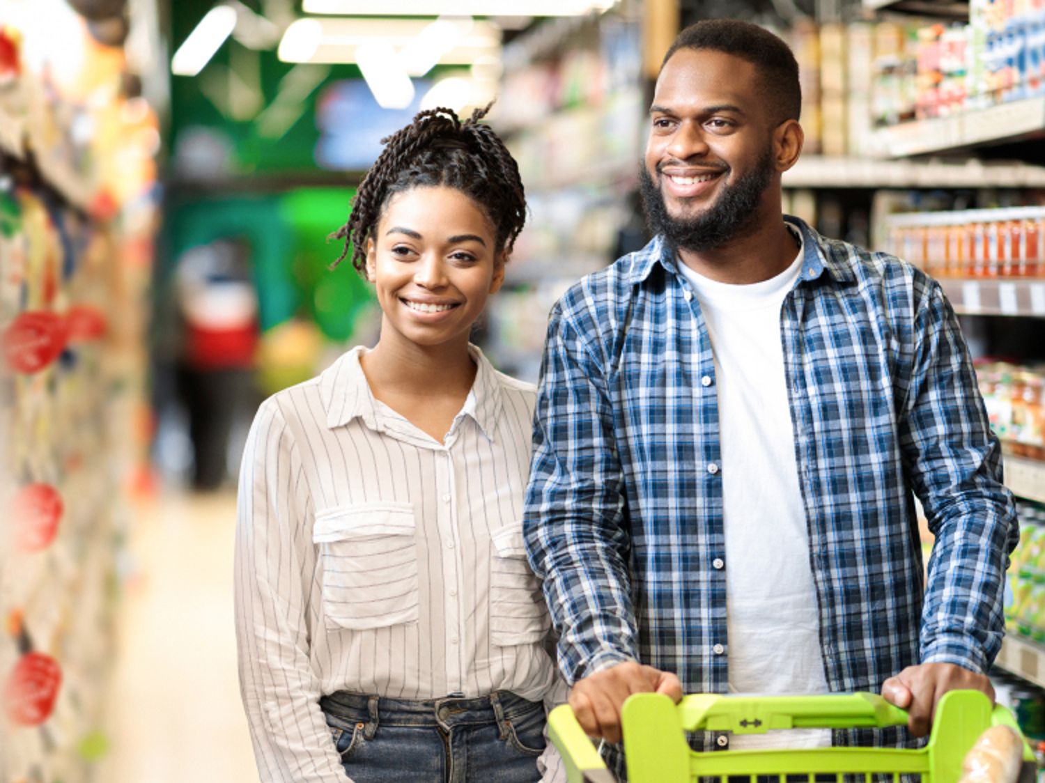 Smiling adult couple shopping together in a grocery store aisle, pushing a cart filled with items.