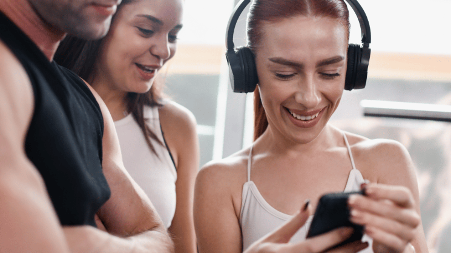 Un grupo de tres personas en el gimnasio, están en forma, una de ellas es una mujer con unos auriculares que sonríe y muestra algo en su teléfono a los demás.